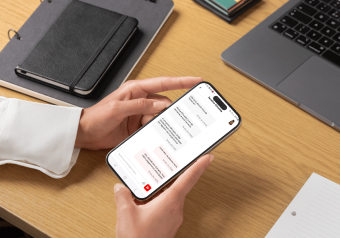 Mãos segurando um smartphone com interface de aplicativo aberta sobre uma mesa com laptop e caderno.