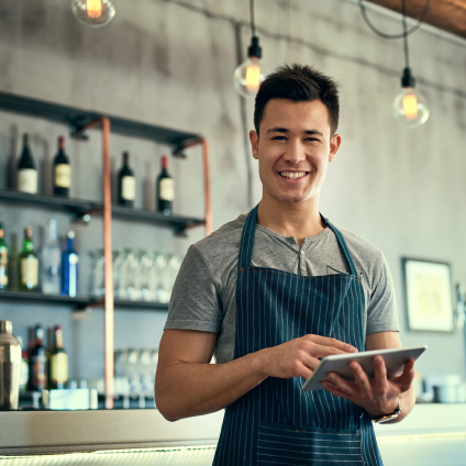 Atendente de bar ou café segurando um tablet atrás do balcão, utilizando tecnologia para gerenciar pedidos e estoque.